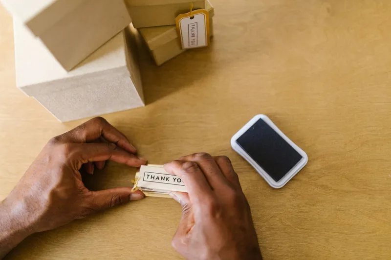 Hands tying a thank-you tag to a gift box beside stacked shipping boxes on a wooden table