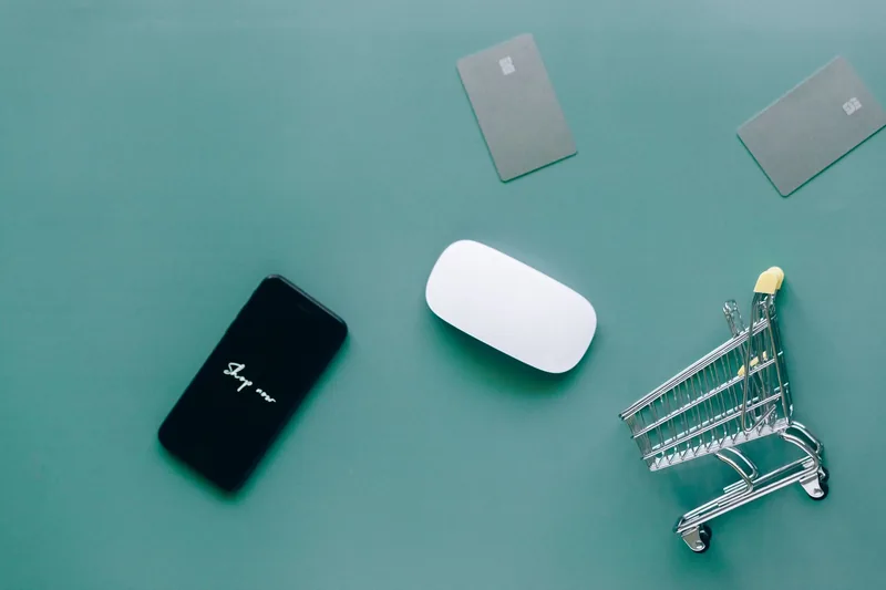 Smartphone, computer mouse, two payment cards and a miniature shopping cart on a green desk
