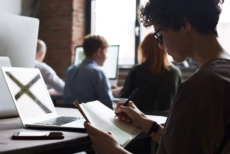 Person taking handwritten notes beside an open laptop in a shared office