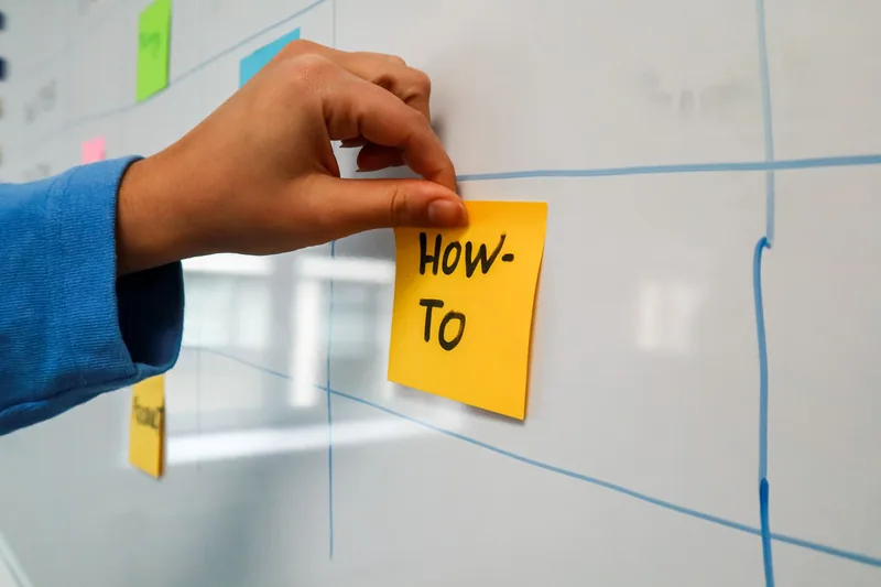 Hand placing a sticky note reading how-to onto a whiteboard content plan