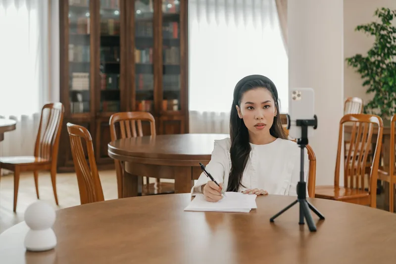 Woman filming a short video on a smartphone mounted on a tripod while taking notes