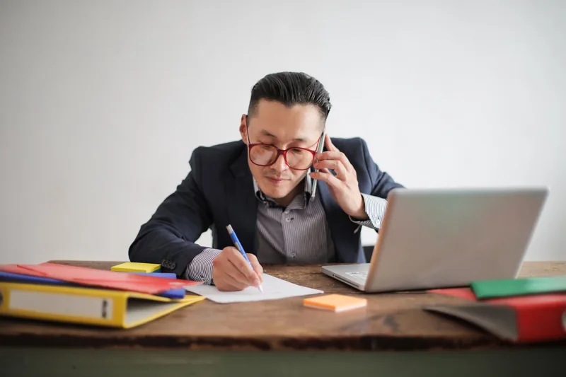 Business owner in a blazer taking notes during a phone call at a desk with a laptop