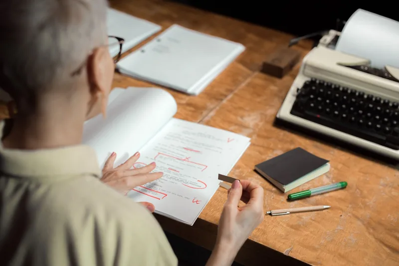 A woman marking up a printed draft with red pen corrections at a desk beside a typewriter