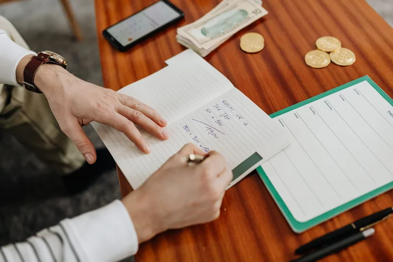 Business owner writing out budget figures in a notebook beside cash and a phone