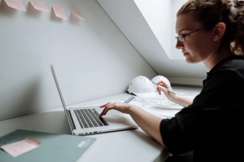 Contractor reviewing plans and job costs on a laptop with a hard hat on the desk