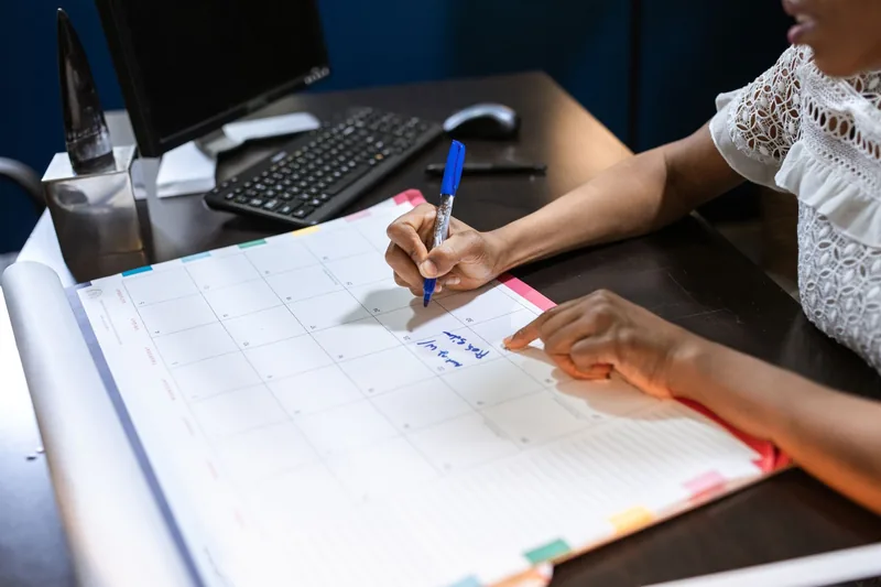 Person writing on a large desk calendar with a blue marker, planning out the month