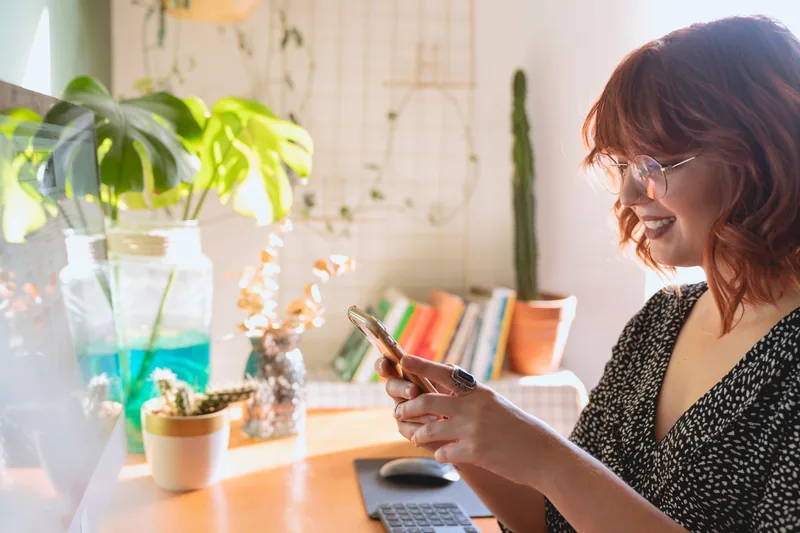 Smiling woman at a desk with plants using her smartphone