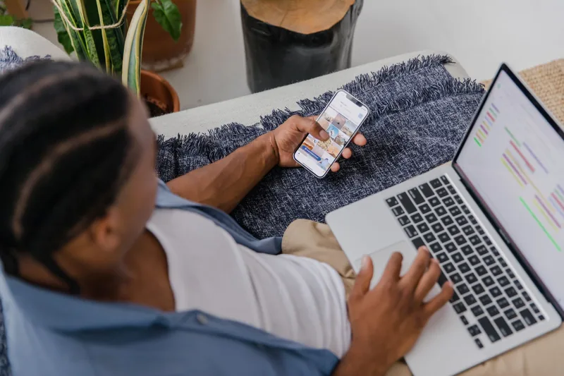 Man on a couch checking a website on his phone with a laptop open beside him