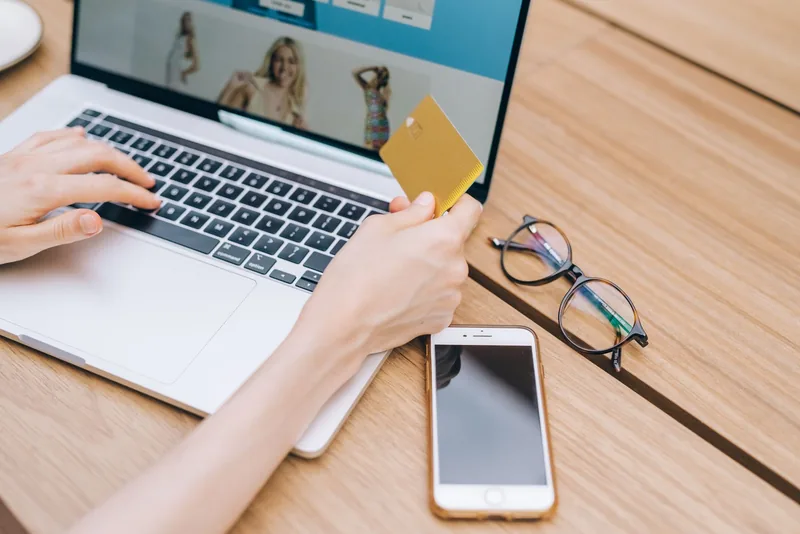Person holding a bank card while shopping on a laptop, with a phone on the desk