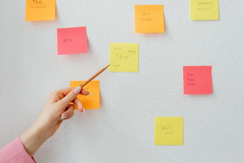 Hand pointing a pen at sticky notes on a wall listing marketing ideas like ads, SEM and web design